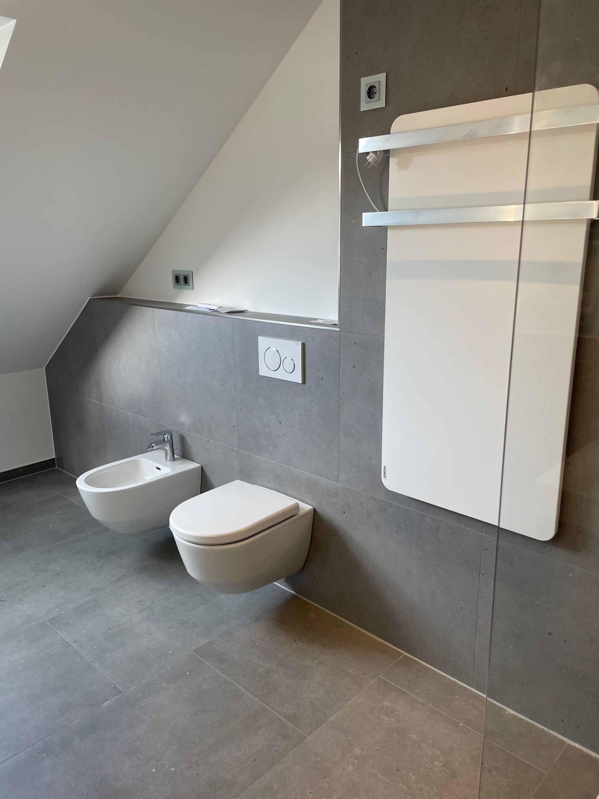 Modernes Badezimmer mit weißem Bidet, WC und Handtuchwärmer an grauer Wand; schräge Decke.