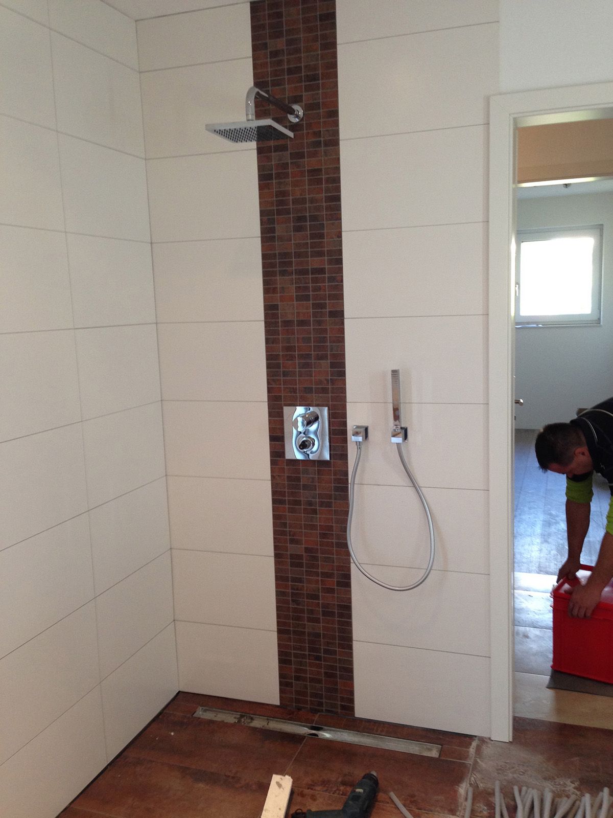 Dusche mit weißen Fliesen, einem braunen Mosaikakzent und einer Person im Türrahmen.