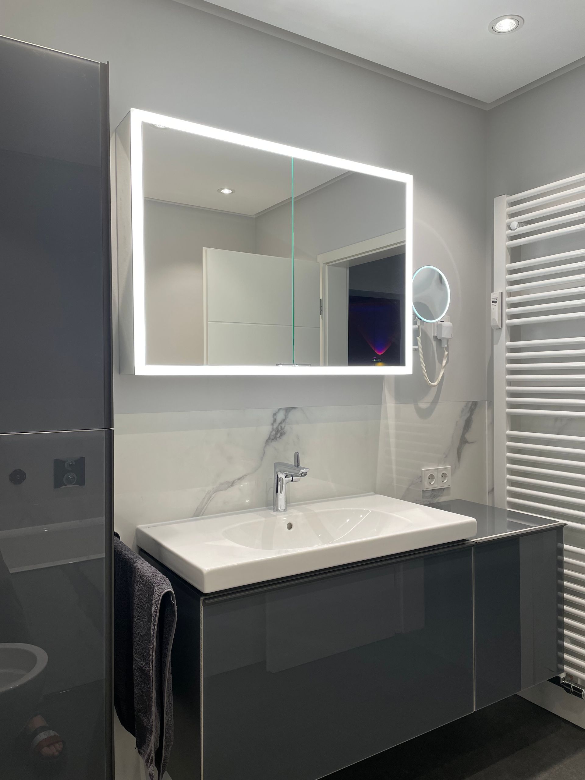 Modernes Badezimmer mit beleuchtetem Spiegel über dem Waschbecken und dunkelgrauem Schrank.