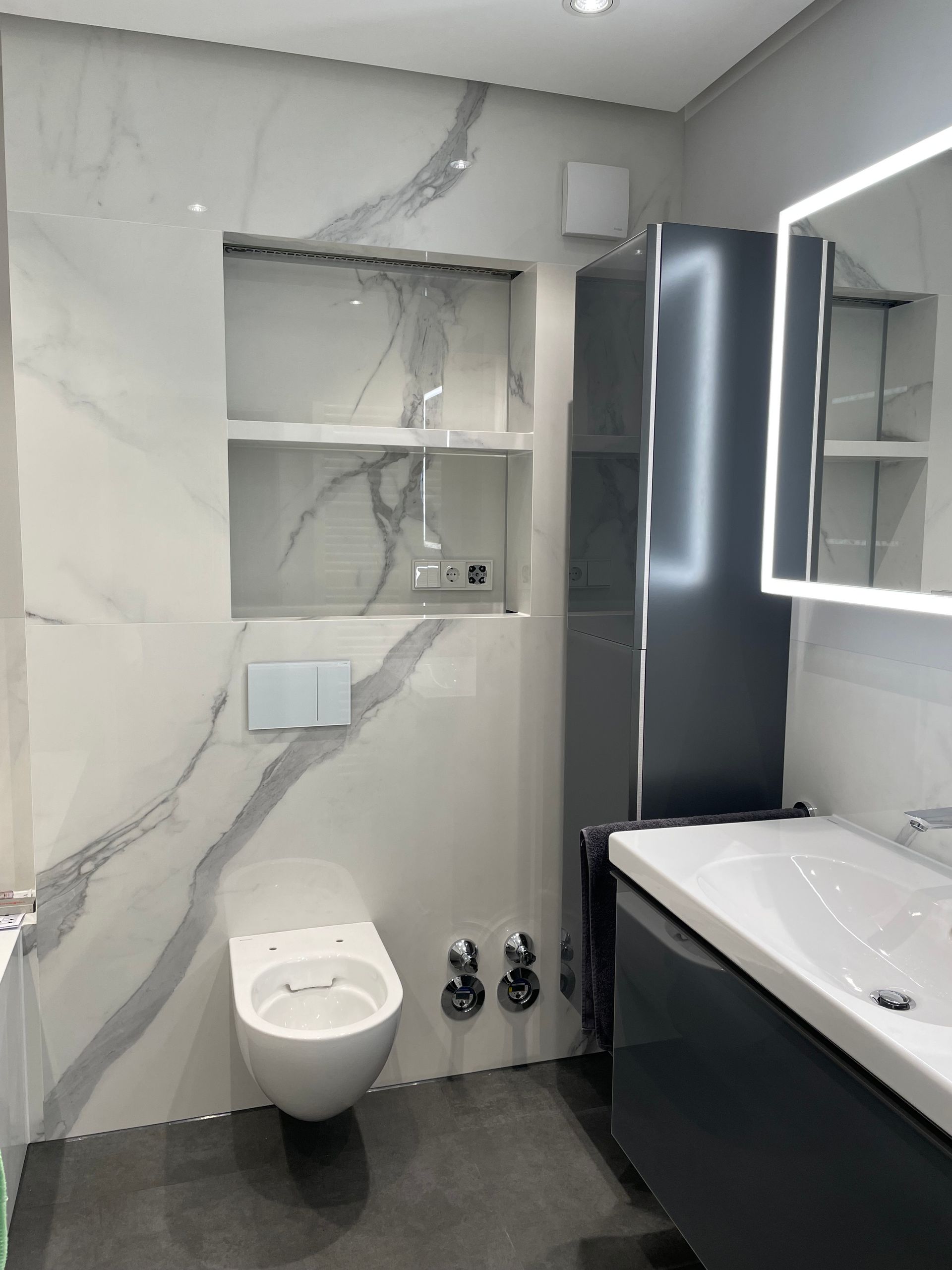 Modernes Badezimmer mit weißen und grauen Fliesen in Marmoroptik, wandhängendem WC und dunkelgrauem Waschtisch.