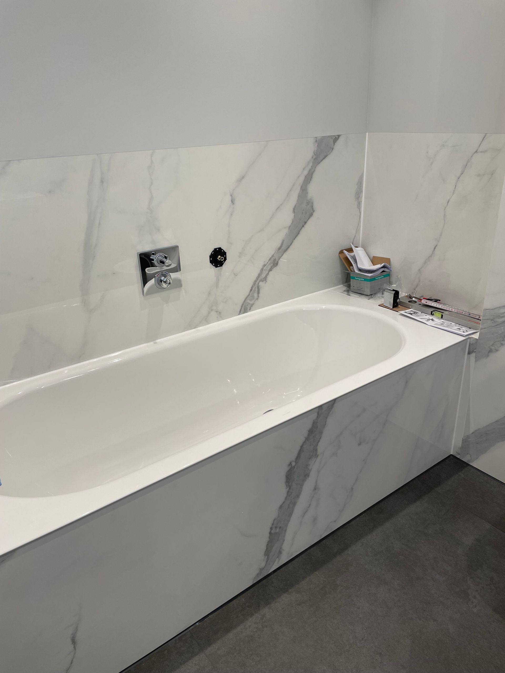 Weiße Badewanne in eine Wand in Marmoroptik eingelassen. Verchromte Armatur, Toilettenartikel und grauer Boden sichtbar.