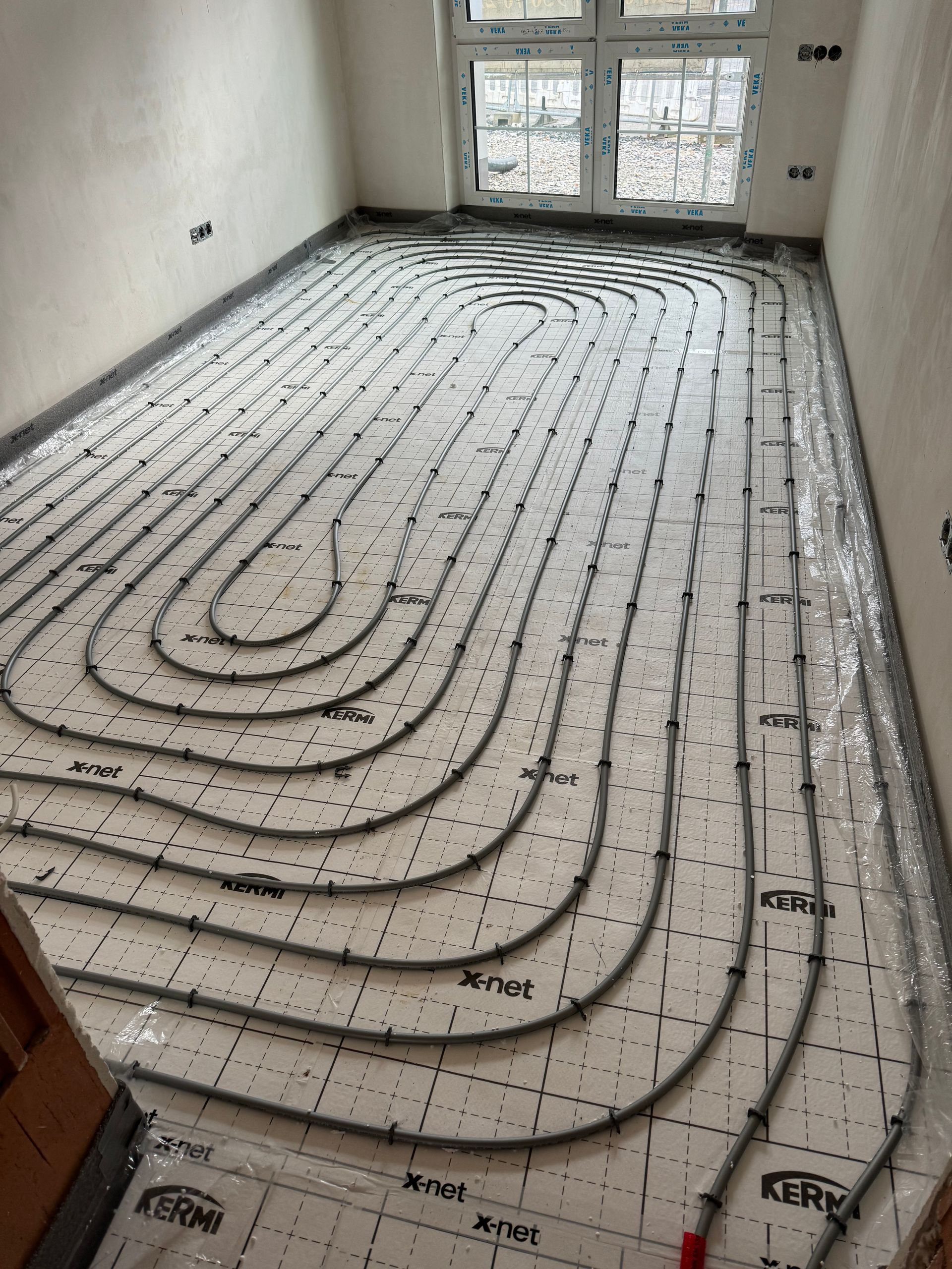 Installation einer Fußbodenheizungsanlage in einem Raum. Graue Rohre verlegt auf weißer Dämmung, in der Nähe eines Fensters.