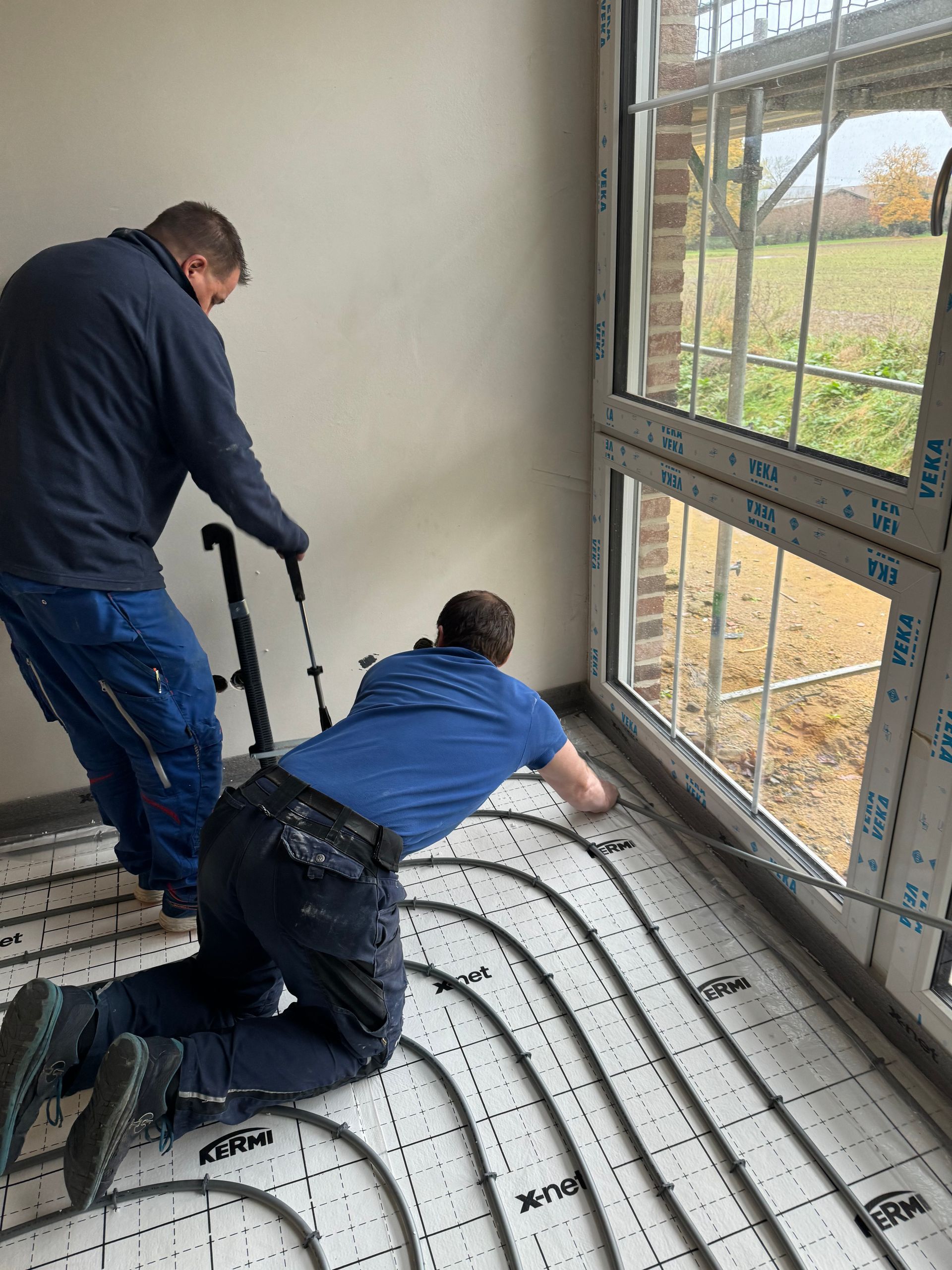 Zwei Arbeiter installieren eine Fußbodenheizung in der Nähe eines großen Fensters, einer kniet, der andere steht.