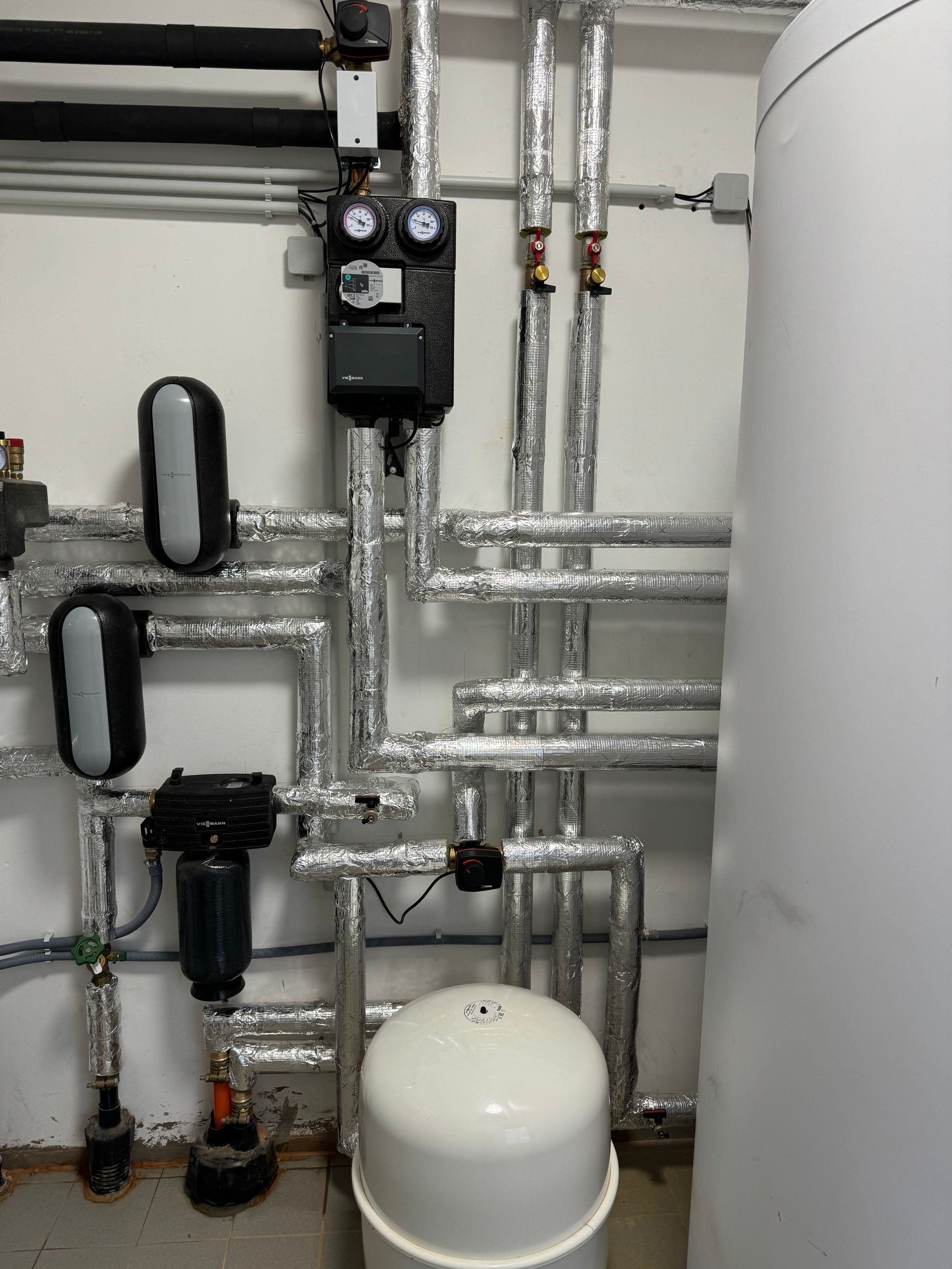 Sanitärsystem mit isolierten Rohren, Pumpen, Manometern und einem weißen Wassertank in einem Hauswirtschaftsraum.