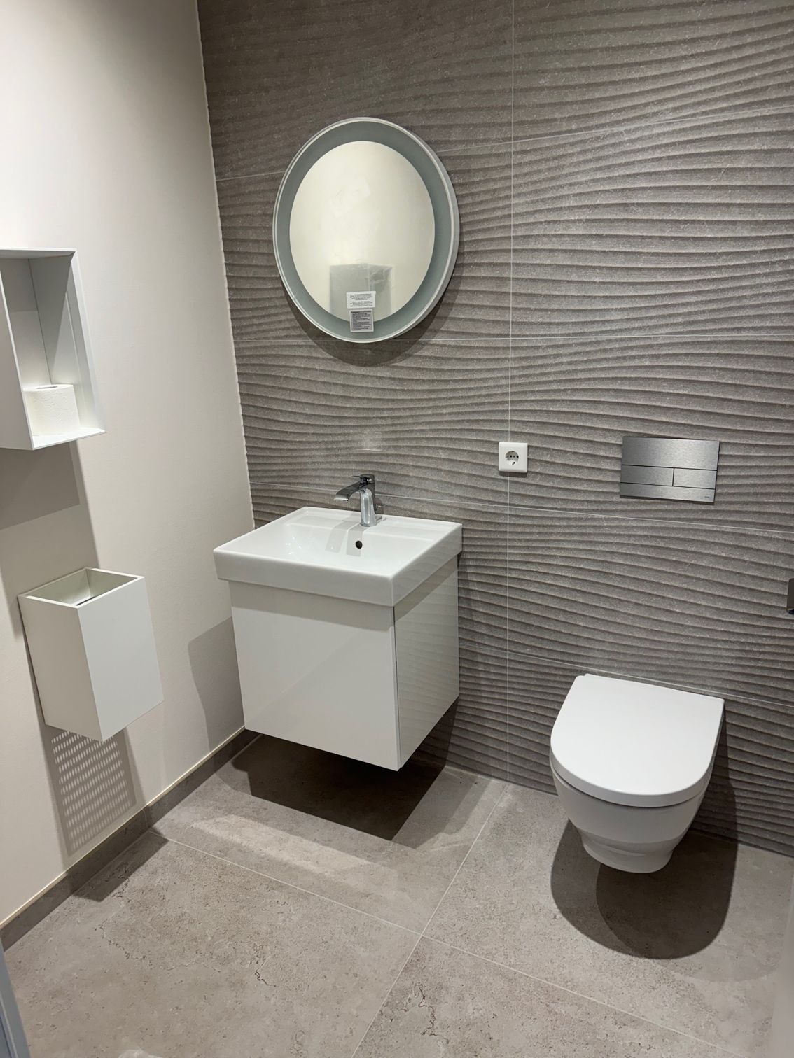 Modernes Badezimmer mit weißem Waschbecken, WC und Stauraum. Graue, strukturierte Wand, runder Spiegel und grauer Boden.