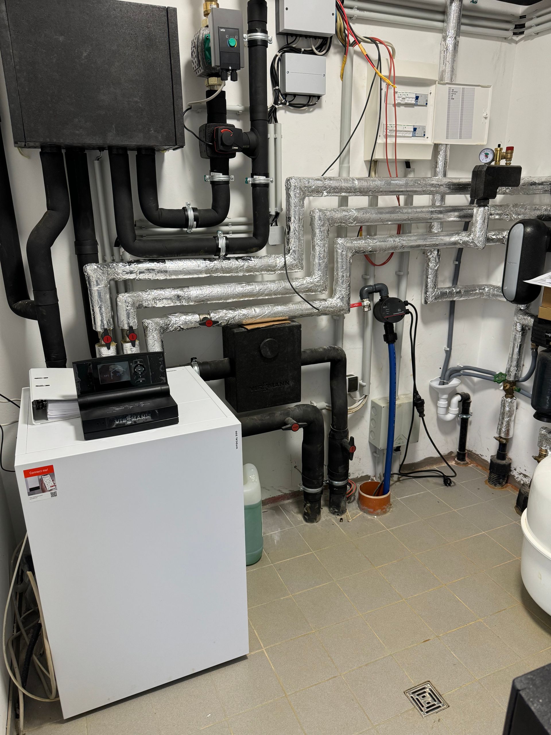 Eine weiße Heizungsanlage mit schwarzen Rohren und Isolierung in einem Hauswirtschaftsraum.