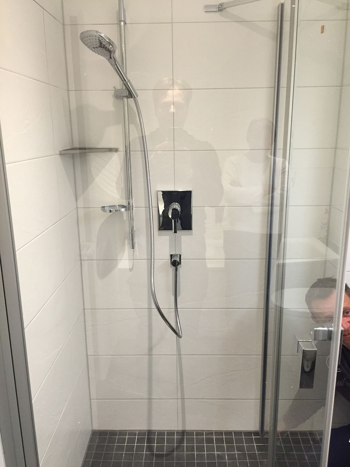 Duschkabine mit Glastür, silbernen Armaturen, weiß gefliesten Wänden und dunkelgrauem Boden.
