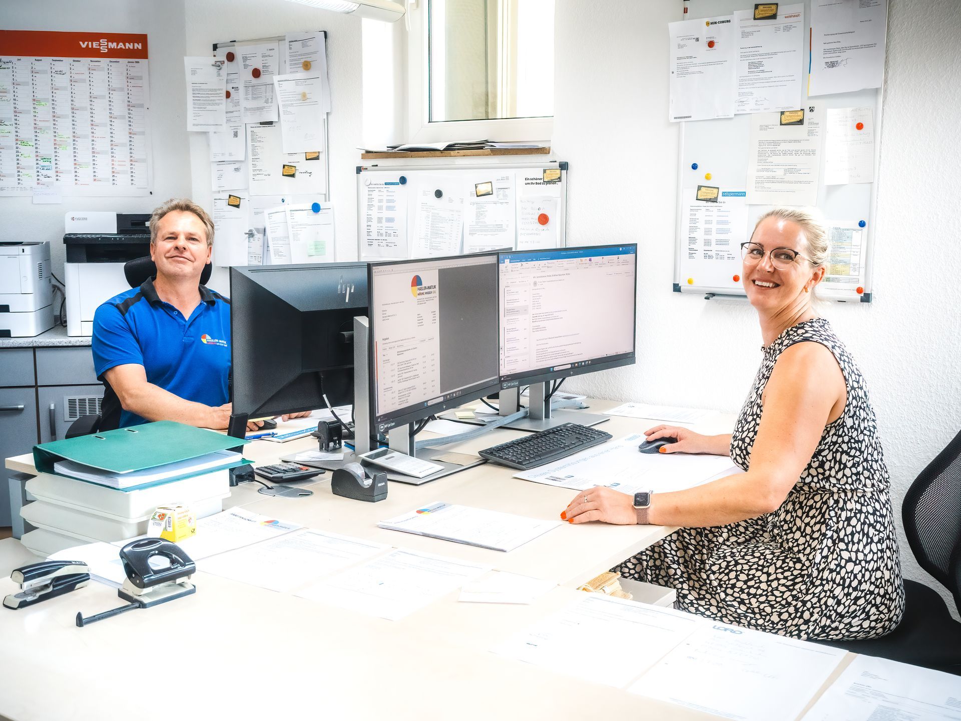 Zwei lächelnde Menschen an einem Schreibtisch mit zwei Computerbildschirmen in einem Büro.
