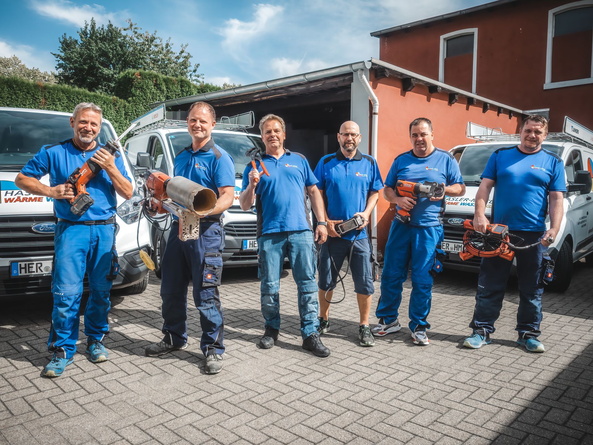 Eine Gruppe von Menschen in blauen Hemden steht mit Lieferwagen vor einem roten Gebäude und grünem Laub.