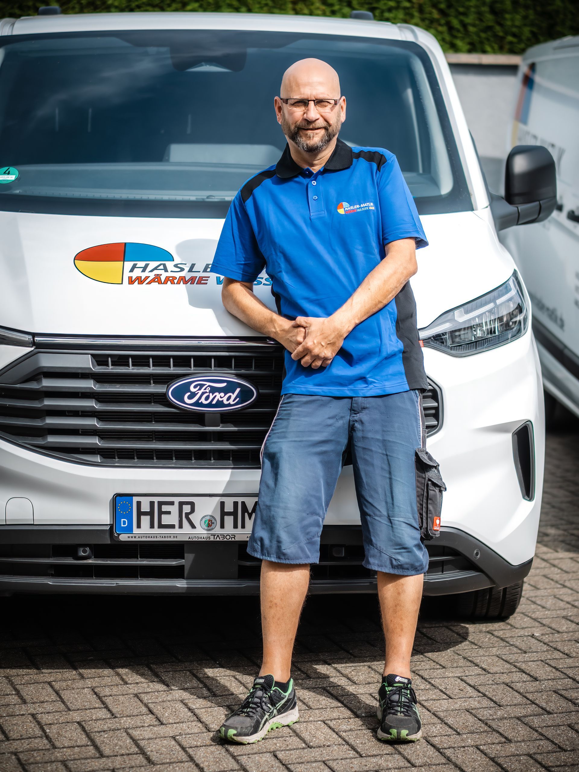 Mann in blauem Hemd und Shorts lehnt mit verschränkten Armen an einem weißen Ford-Van. Nummernschild „HER HM“.
