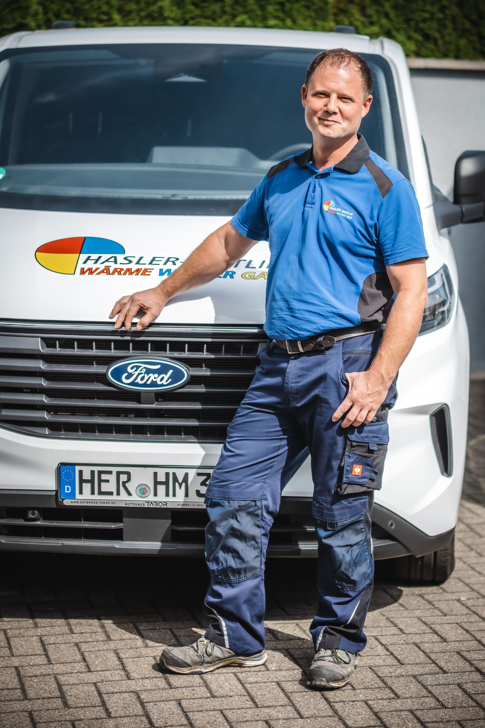 Mann in Arbeitskleidung steht neben einem weißen Ford-Transporter mit Firmenlogo. Außenaufnahme.