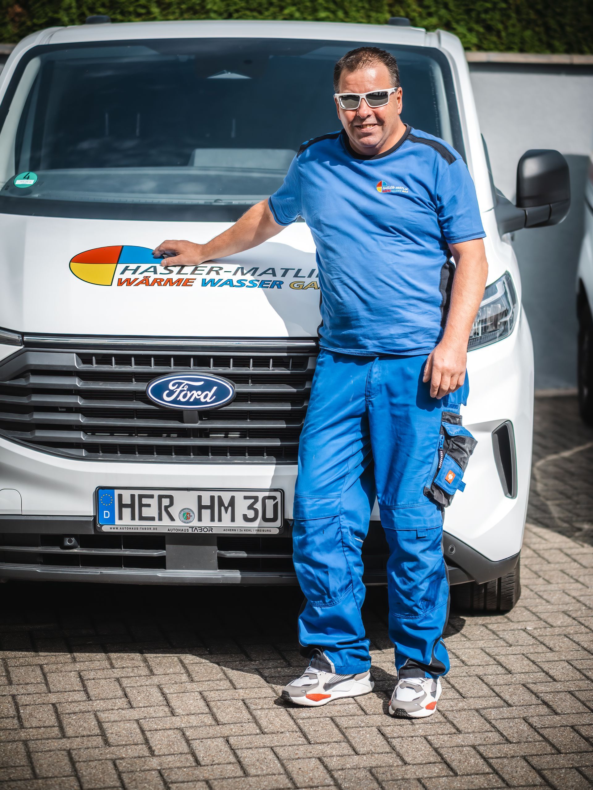 Ein Mann in blauer Arbeitskleidung steht neben einem weißen Ford-Transporter mit Firmenlogo.