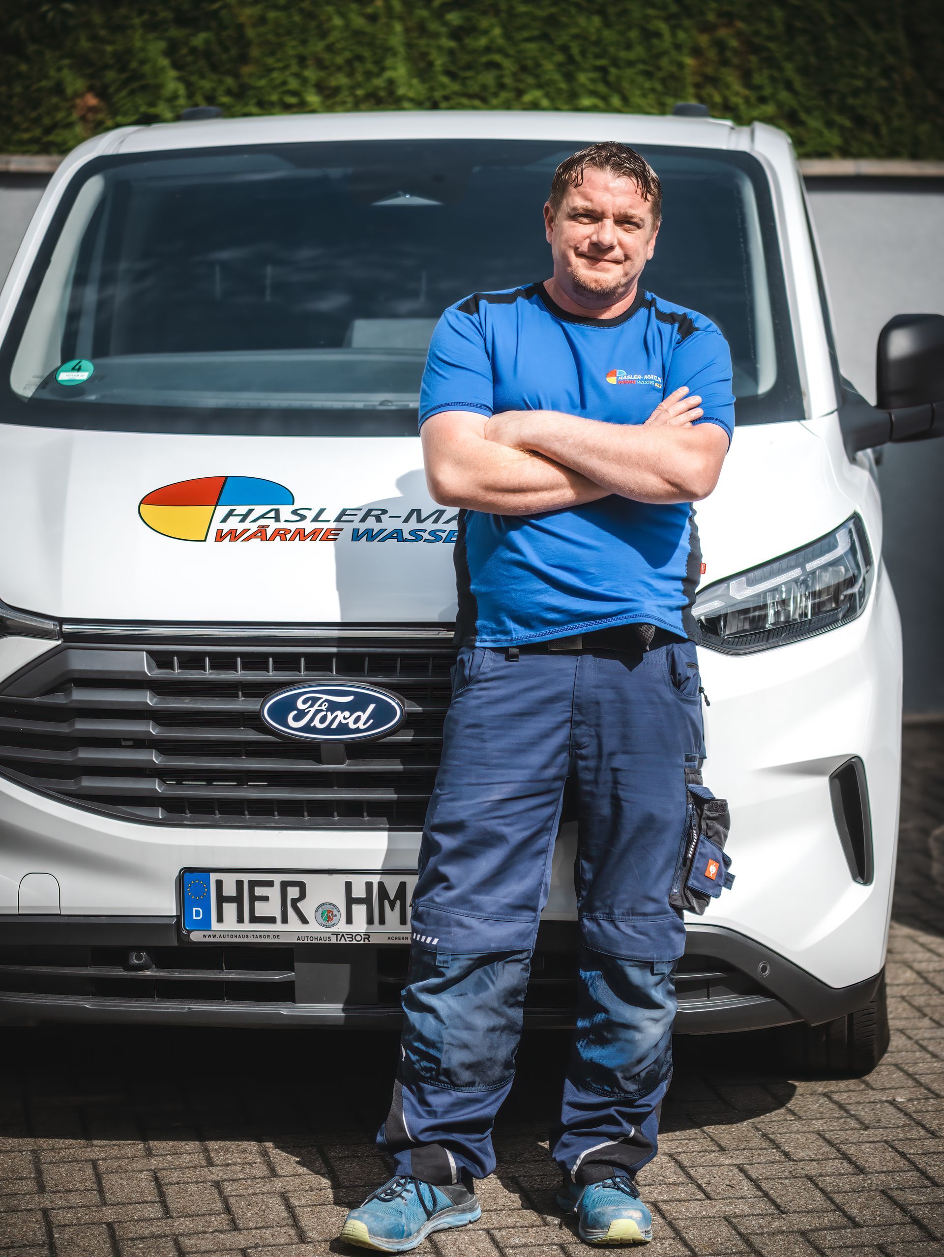 Mann in blauer Uniform mit verschränkten Armen vor einem weißen Ford-Van. Nummernschild „HER-HM“.
