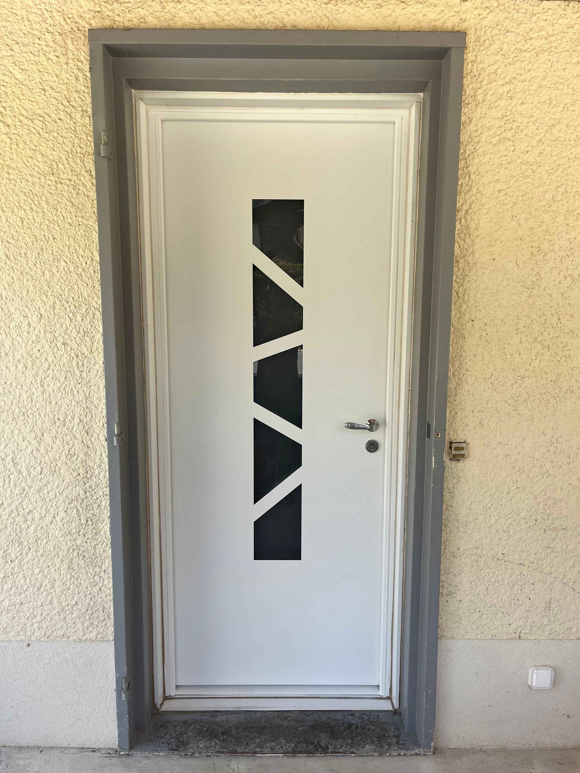Porte d'entrée blanche avec un panneau de verre géométrique étroit intégré à un mur en stuc et un cadre gris.
