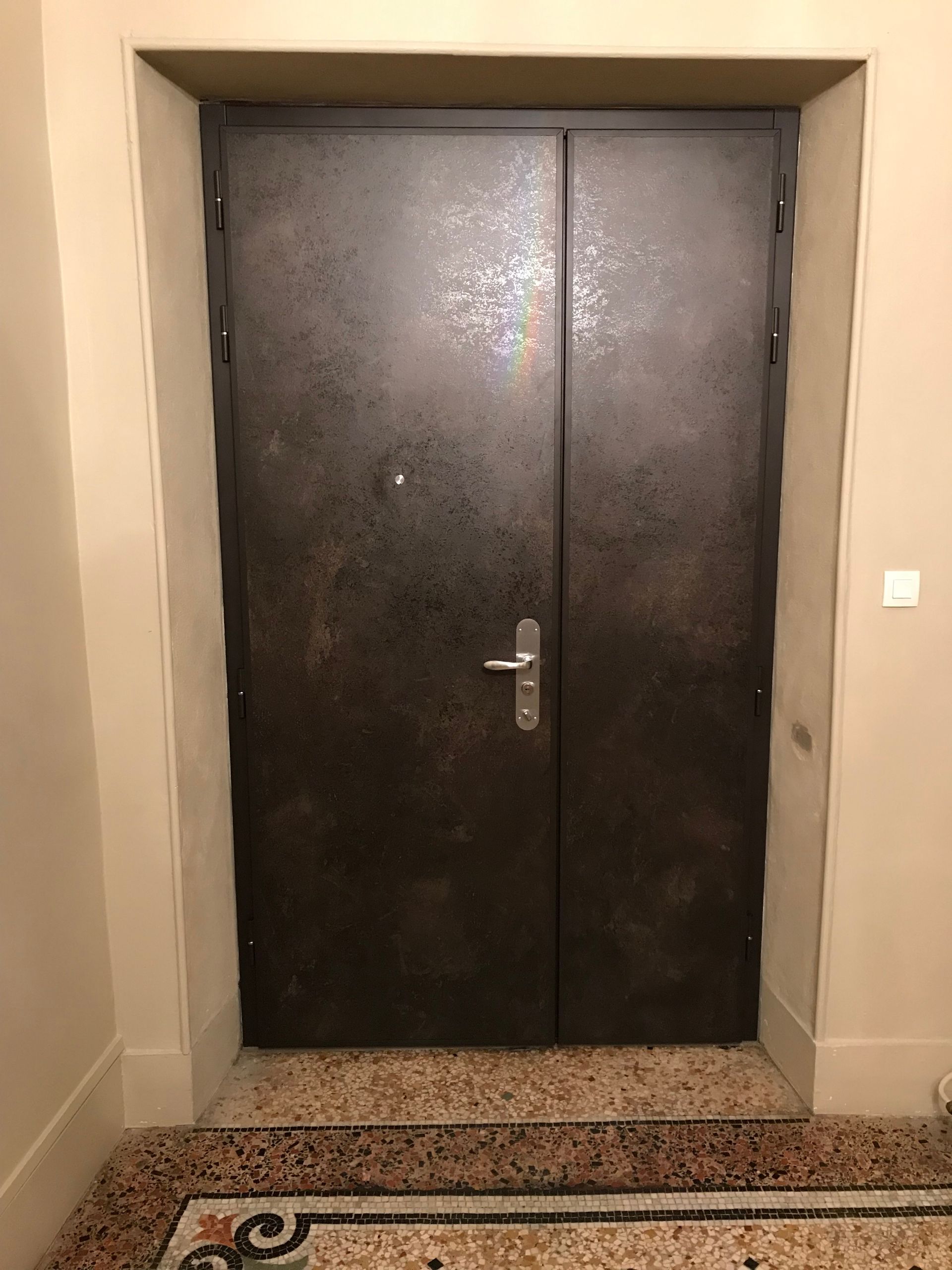 Porte double en métal foncé avec poignée, dans un cadre de porte beige, avec un sol décoratif.