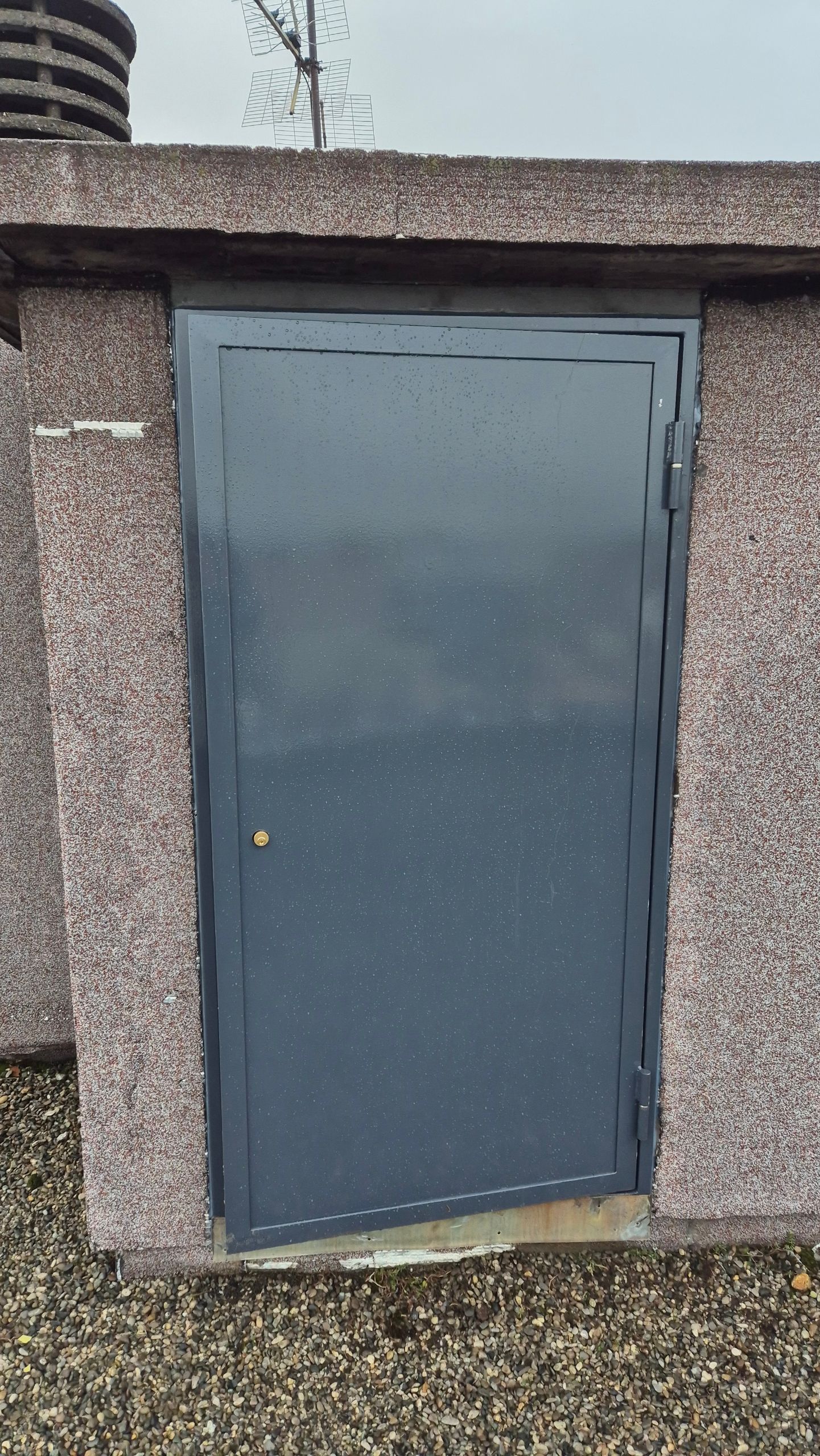 Porte en métal gris encastrée dans un mur en béton texturé, avec une petite poignée de porte en laiton.