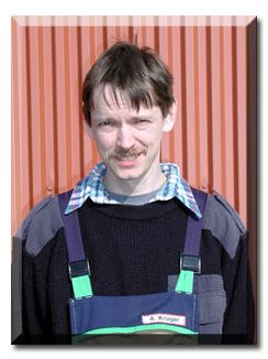Ein Mann in Overall und Pullover lächelt vor einer roten Wand.