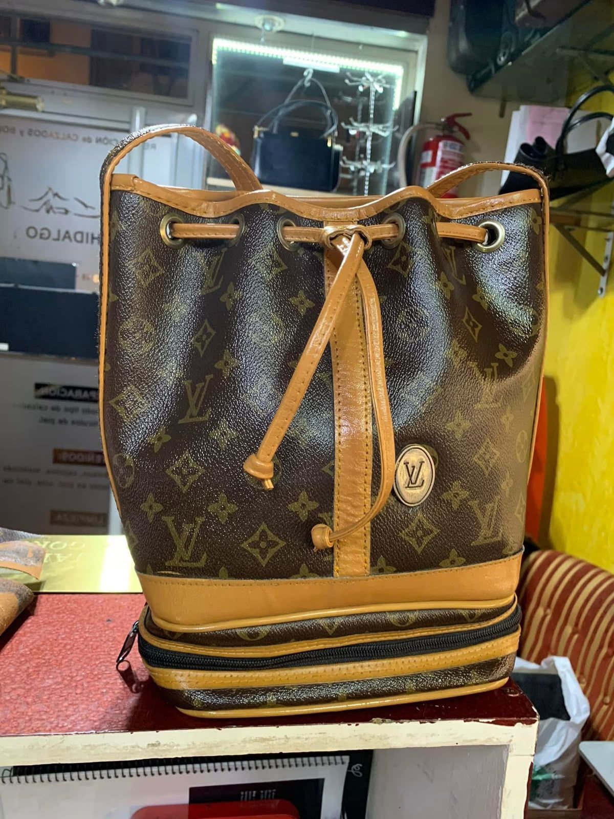 Bolso marrón con monograma, cordón ajustable, ribetes color tostado y base a rayas, sobre una estantería de exposición