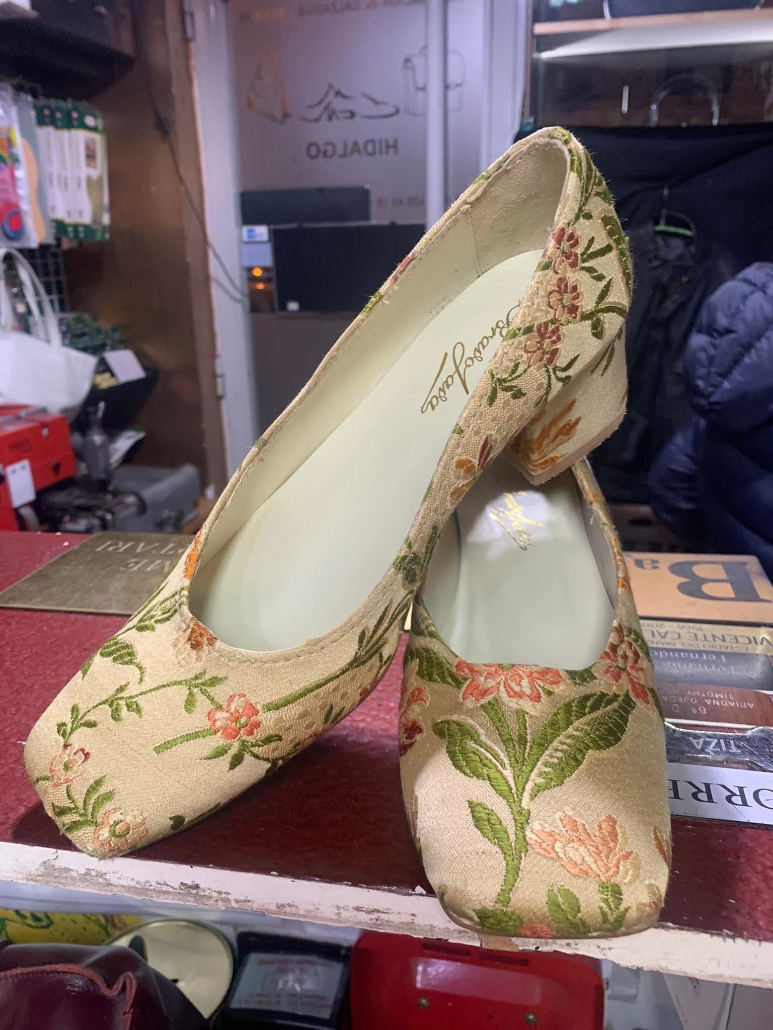 Un par de zapatos de tacón beige para mujer con bordados florales en un estante, uno de ellos colocado en posición vertical.