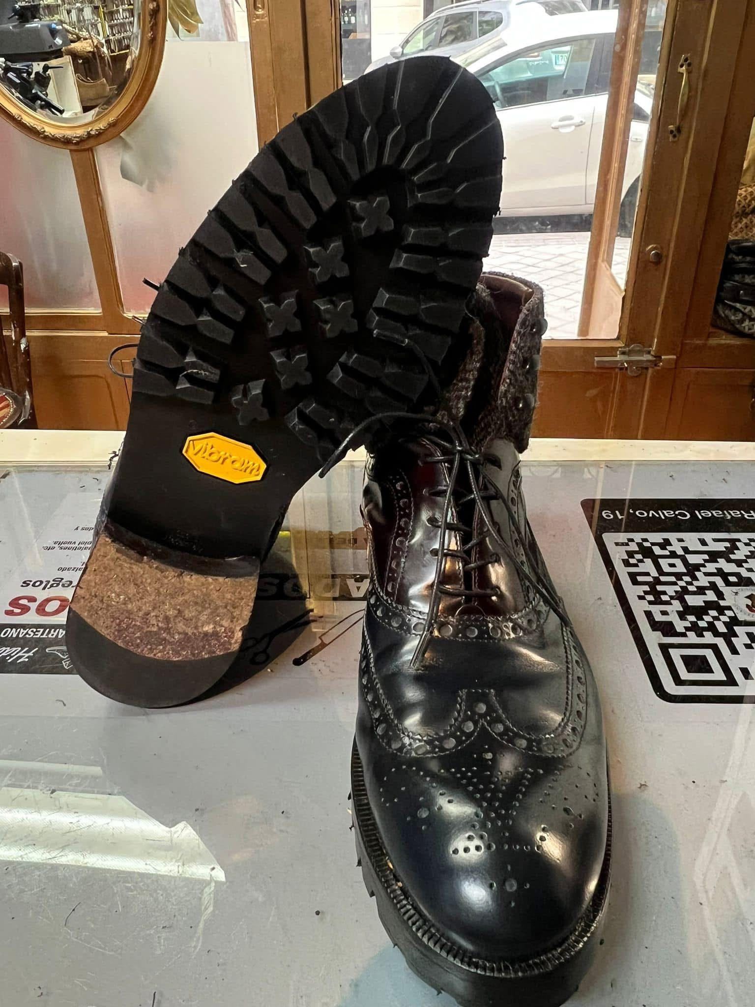 Zapatos de vestir de cuero negro en exhibición, uno en posición vertical y el otro mostrando la suela con relieve, sobre una superficie estampada.
