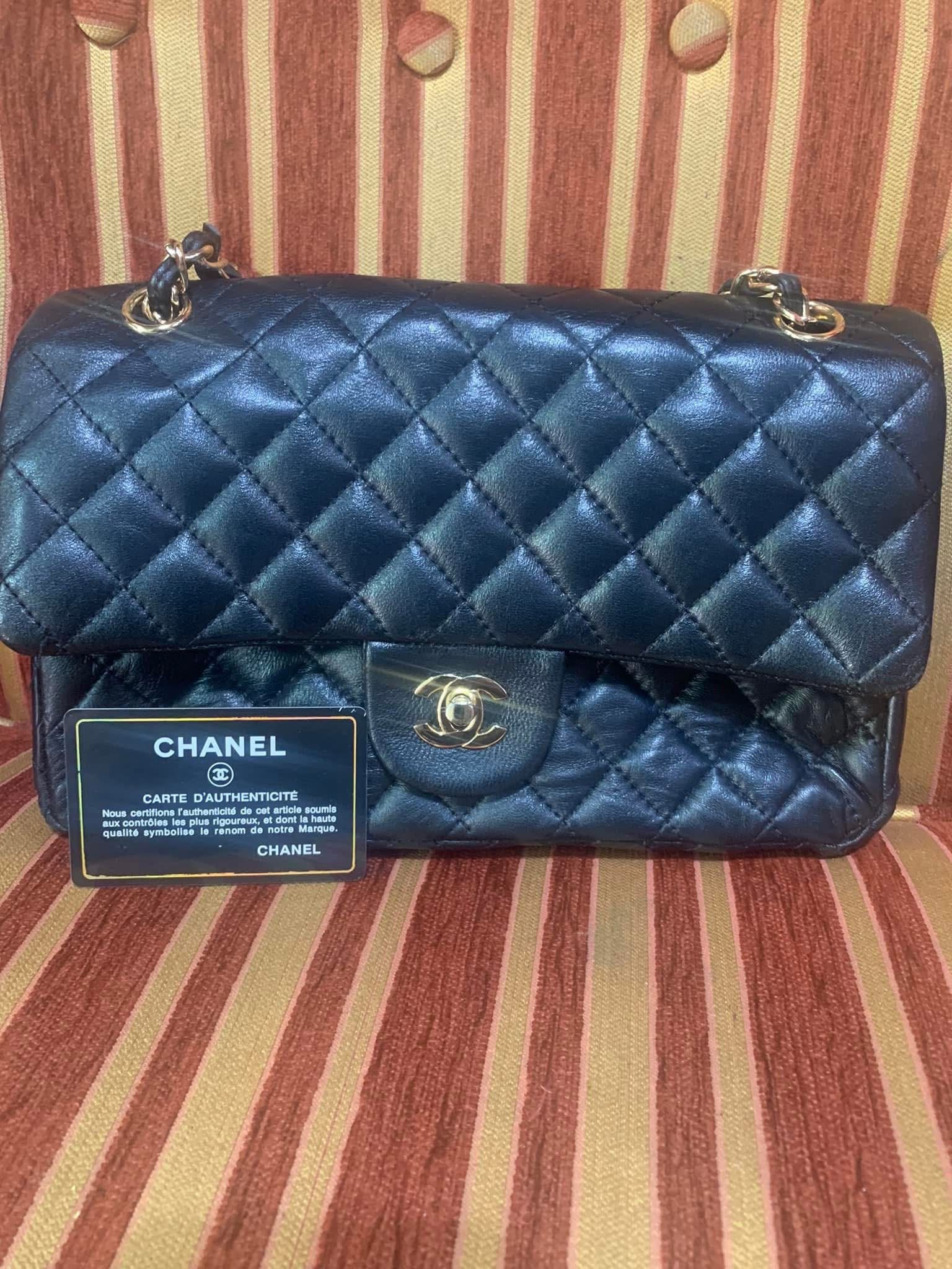 Bolso Chanel negro acolchado con cadena dorada sobre una silla a rayas, con una etiqueta Chanel colgando