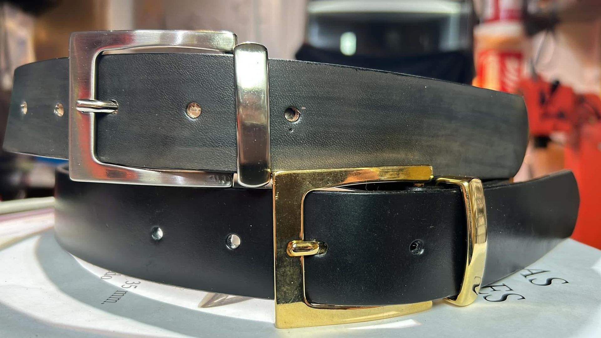 Dos cinturones de cuero negro, uno con hebilla plateada y otro con hebilla dorada, apilados sobre una superficie de exposición