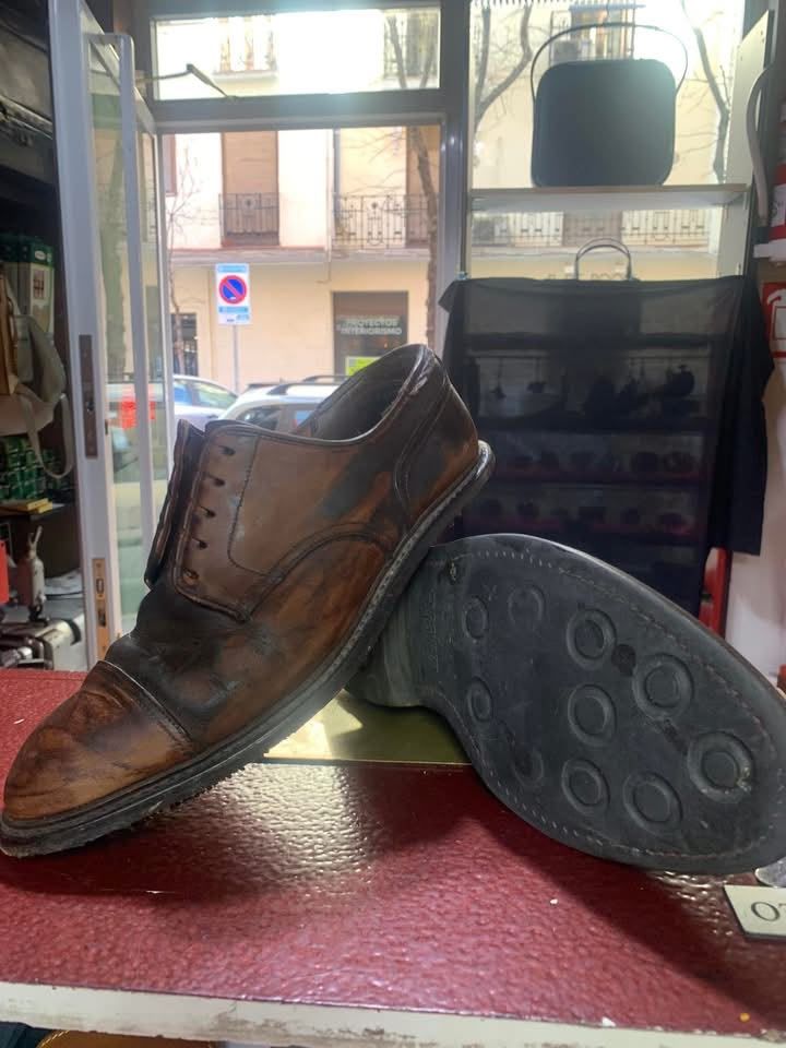 Un zapato de cuero marrón desgastado sobre el mostrador de una tienda, junto a otro zapato, con una vista de la calle al fondo