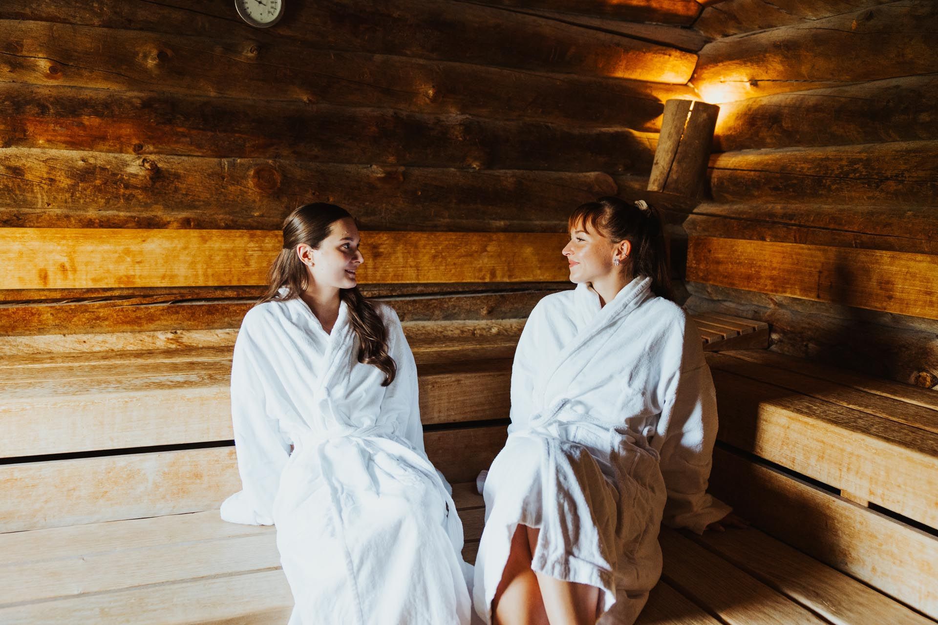 Zwei Personen in weißen Gewändern sitzen in einer Holzsauna und schauen einander an.