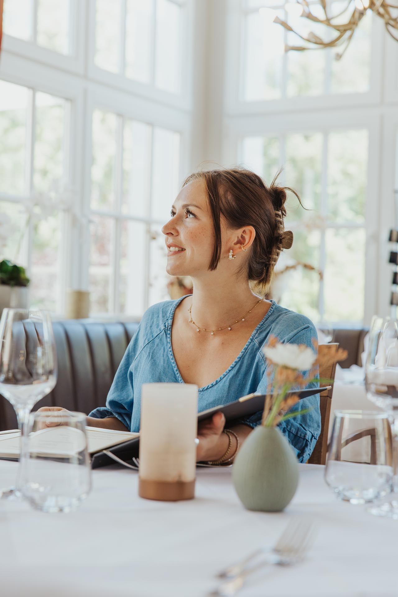 Eine Frau sitzt an einem Restauranttisch, lächelt und blickt nach oben; sie hält einen Notizblock in der Hand.