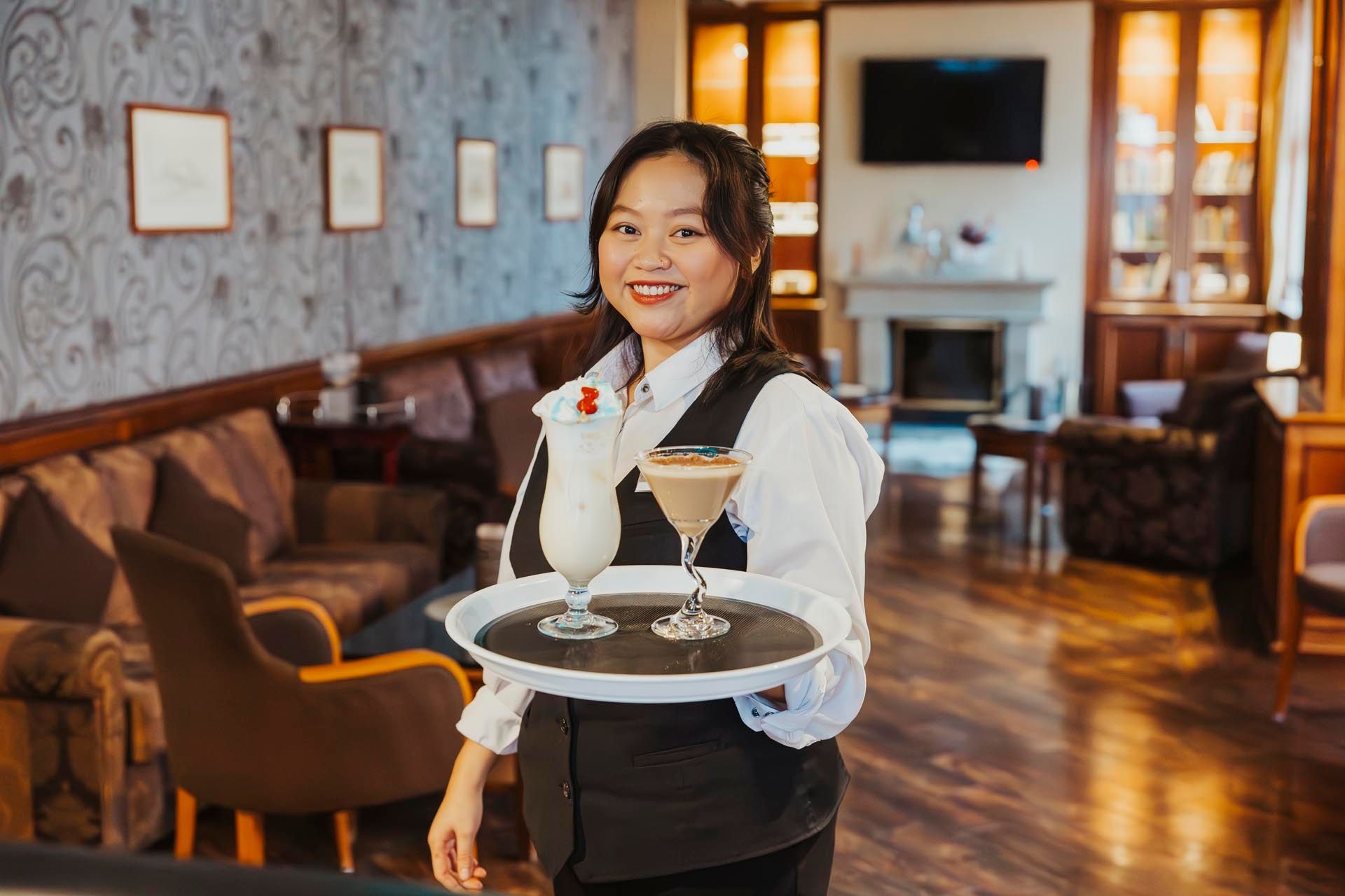 Kellnerin mit Tablett und zwei Cocktails in einer Lounge mit Kamin, lächelnd.