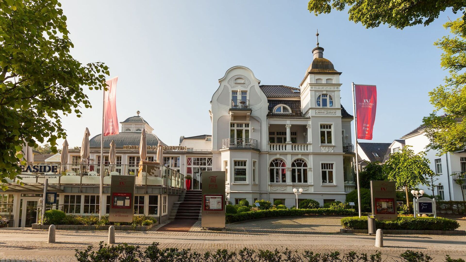 Elegantes weißes Gebäude mit Turm; rote Fahnen, Sitzbereich im Freien und Schilder an einem sonnigen Tag.
