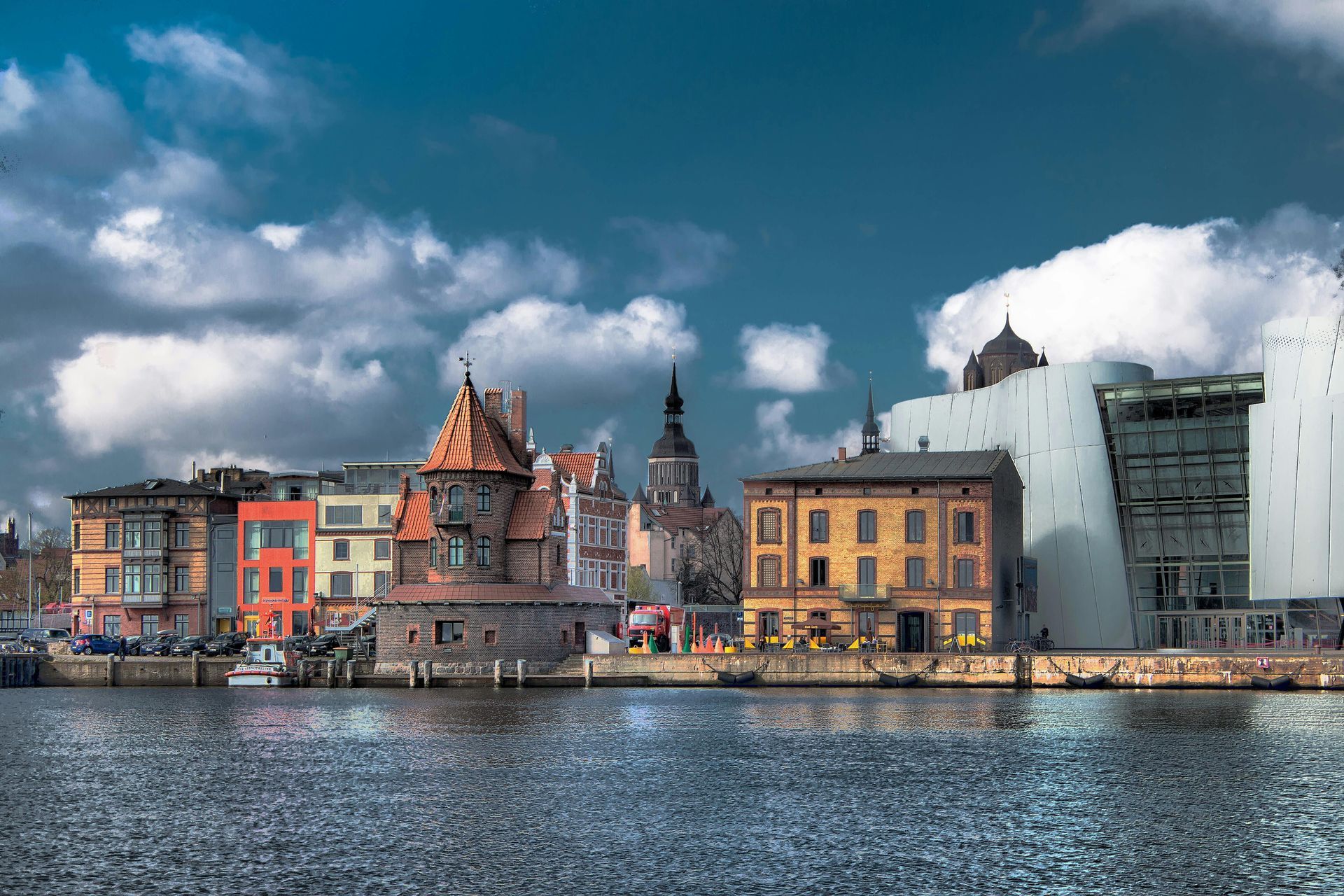 Stadtbild mit farbenfrohen Gebäuden, einem Turm und moderner Architektur an einer Uferpromenade unter bewölktem Himmel.