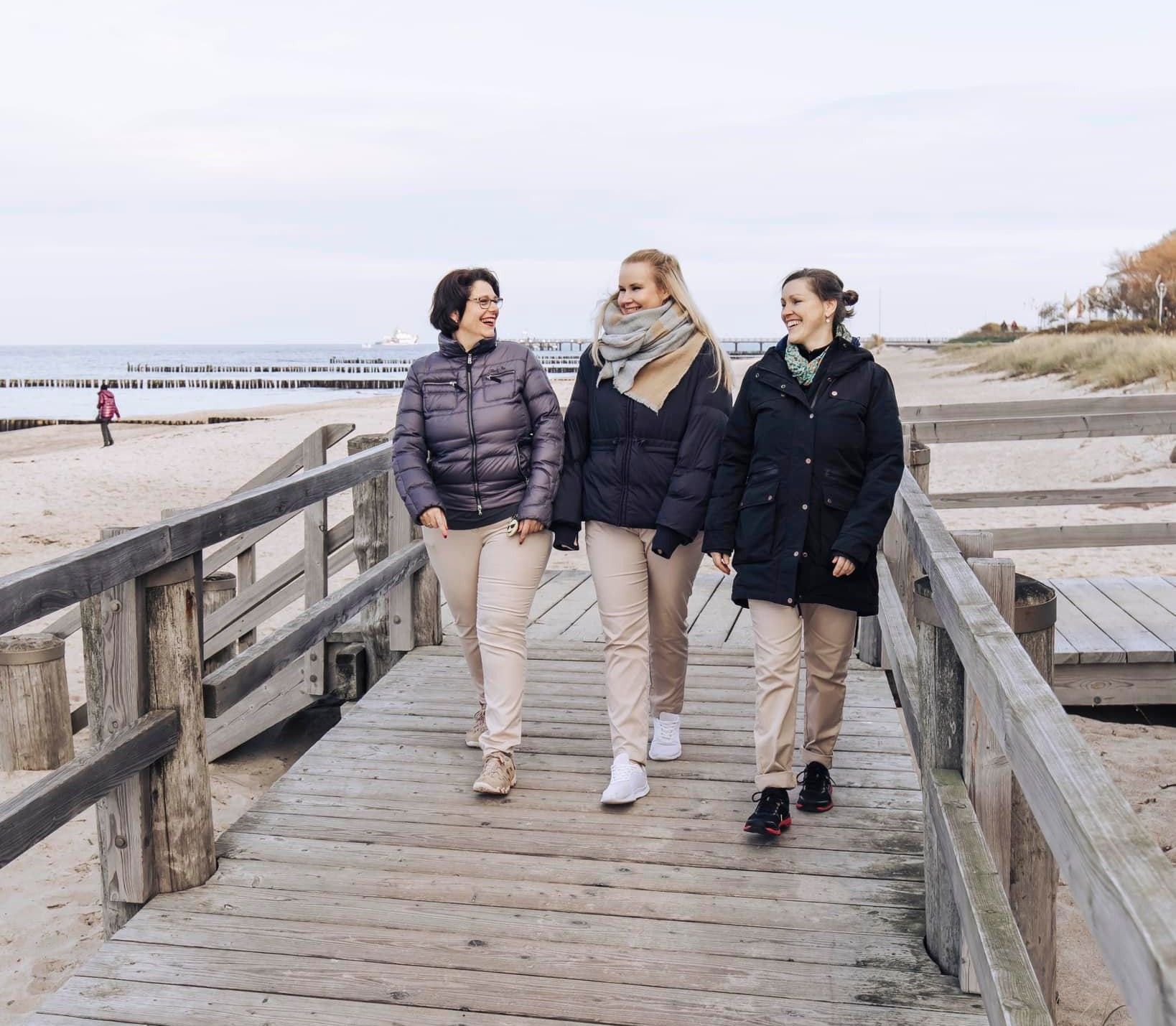 Drei Personen in Wintermänteln und leichten Hosen spazieren und unterhalten sich auf einem Holzsteg an einem Sandstrand.