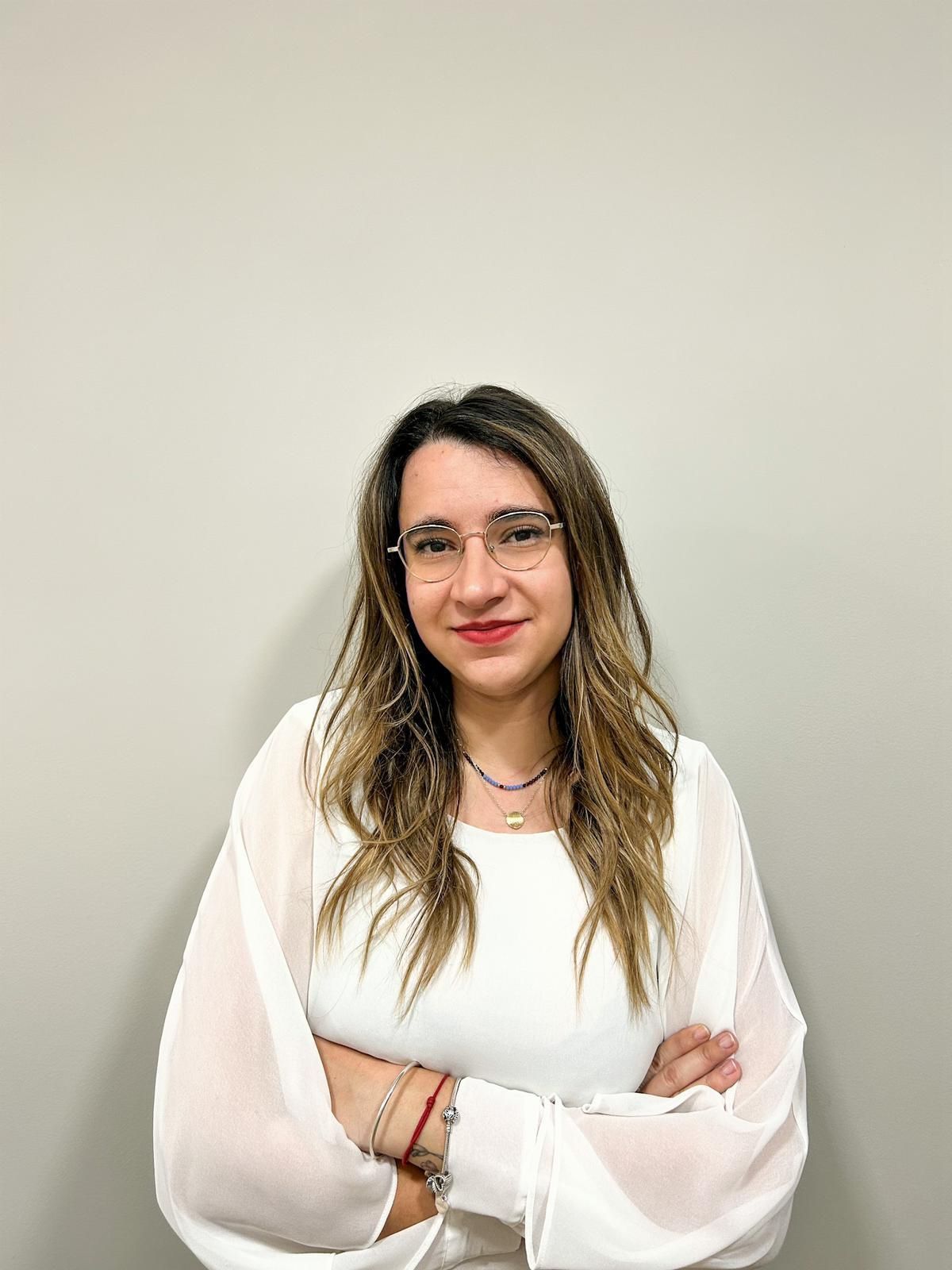 Una mujer que lleva gafas y una camisa blanca está de pie con los brazos cruzados.