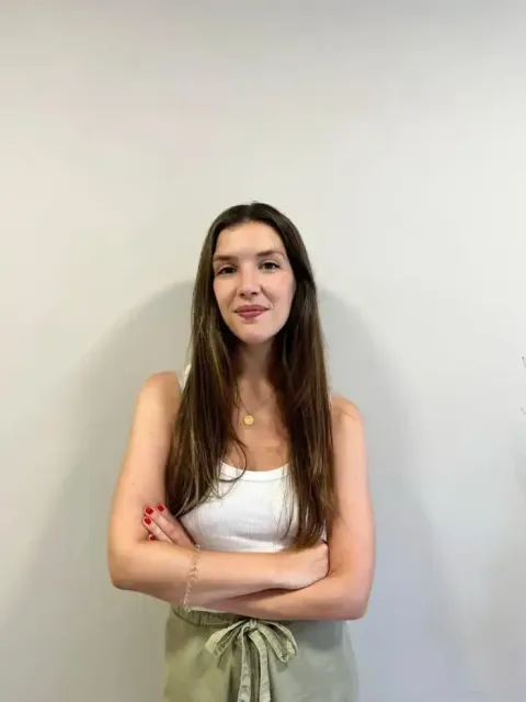 Mujer con los brazos cruzados, vestida con una camiseta blanca y pantalones verdes