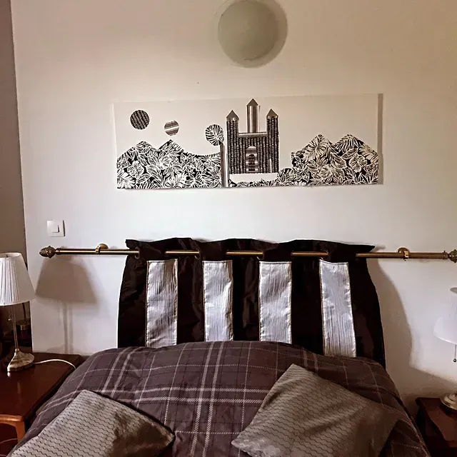 Une chambre avec un lit recouvert d'une couette à carreaux marron et d'une tête de lit façon rideau, sous un tableau représentant un paysage en noir et blanc.