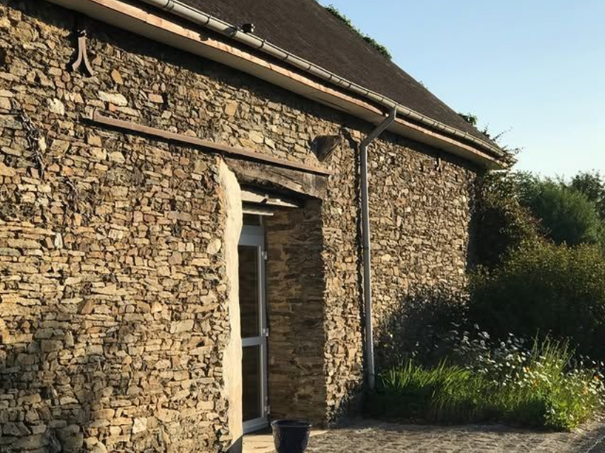 Un bâtiment en pierre au toit sombre et à l'entrée vitrée en retrait, se détachant sur un fond d'arbres et de verdure.
