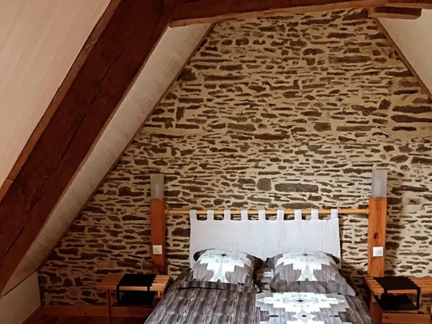 Une chambre avec un lit adossé à un mur de pierre rustique, sous un plafond lambrissé en pente, et une parure de lit à motifs.