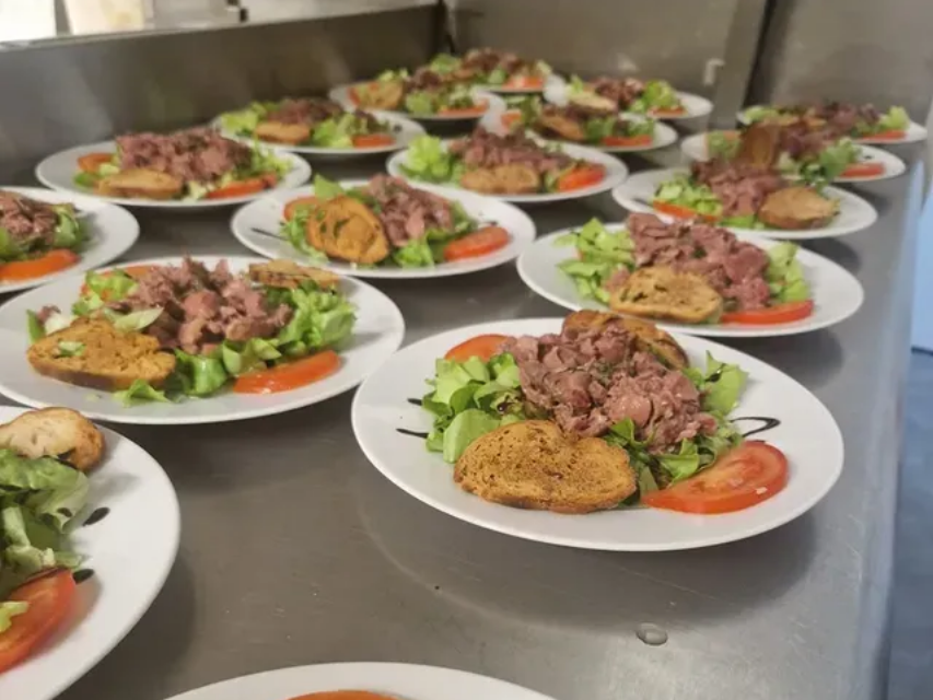 De nombreuses assiettes blanches disposées sur un comptoir en acier inoxydable, chacune contenant une salade avec de la viande, de la verdure, une tranche de tomate et du pain grillé.