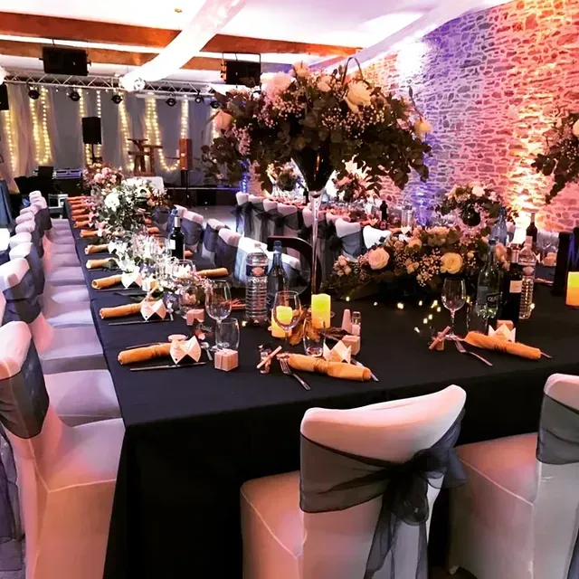 Une longue table de banquet dressée avec des nappes noires, des centres de table floraux et des chaises aux ceintures anthracite, dans une salle en pierre éclairée.