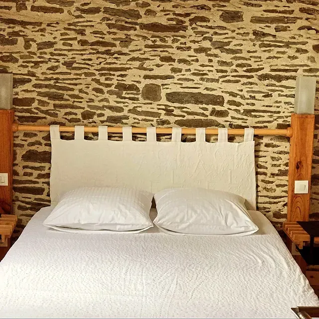 Un lit avec deux oreillers blancs et une tête de lit blanche, adossé à un mur de pierre rustique dans une chambre.
