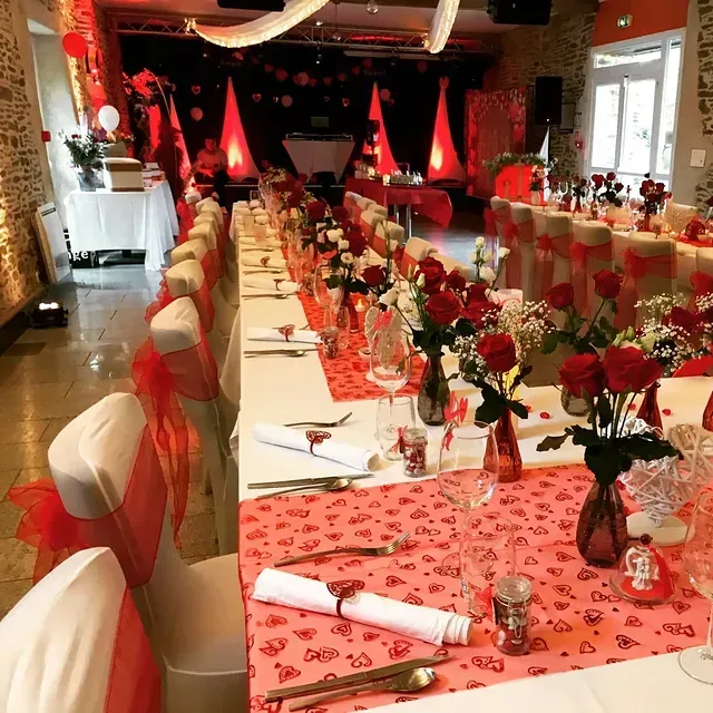 Une salle de banquet décorée pour un événement romantique avec de longues tables, des chemins de table rouges à motifs de cœurs, des roses rouges et des chaises ornées de nœuds rouges.