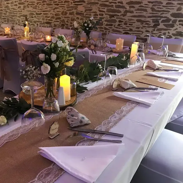 De longues tables de banquet dressées pour un mariage, avec des chemins de table en toile de jute bordés de dentelle, des centres de table floraux et des bougies.