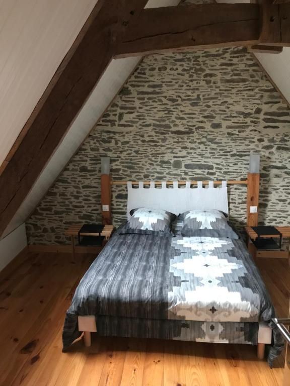 Chambre avec poutres en bois inclinées, mur en pierre, lit double avec couette grise à motifs et tables de chevet en bois sur parquet.
