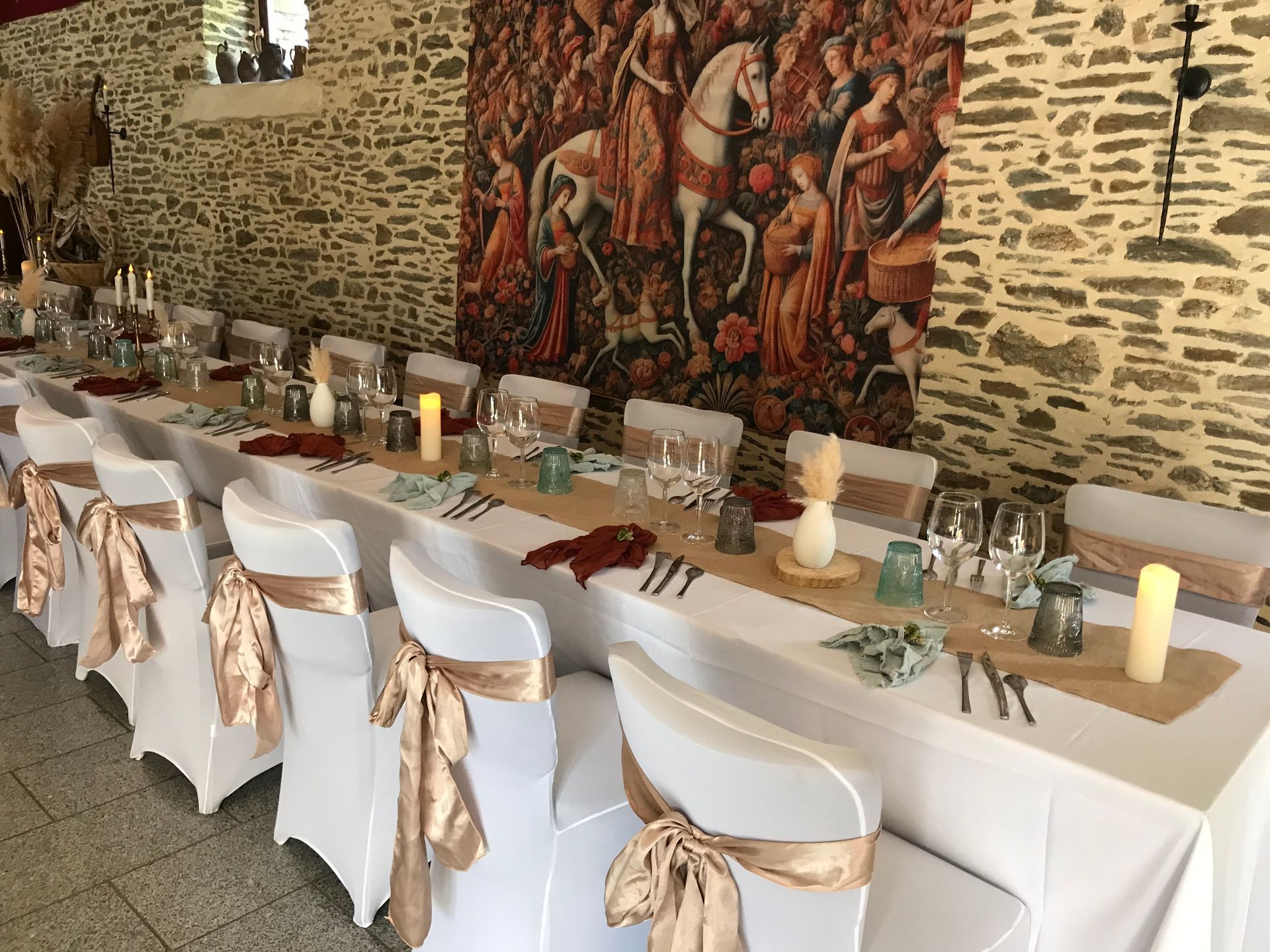 Une longue table est dressée pour un événement formel dans une salle aux murs de pierre ornée d'une grande tapisserie de style médiéval.