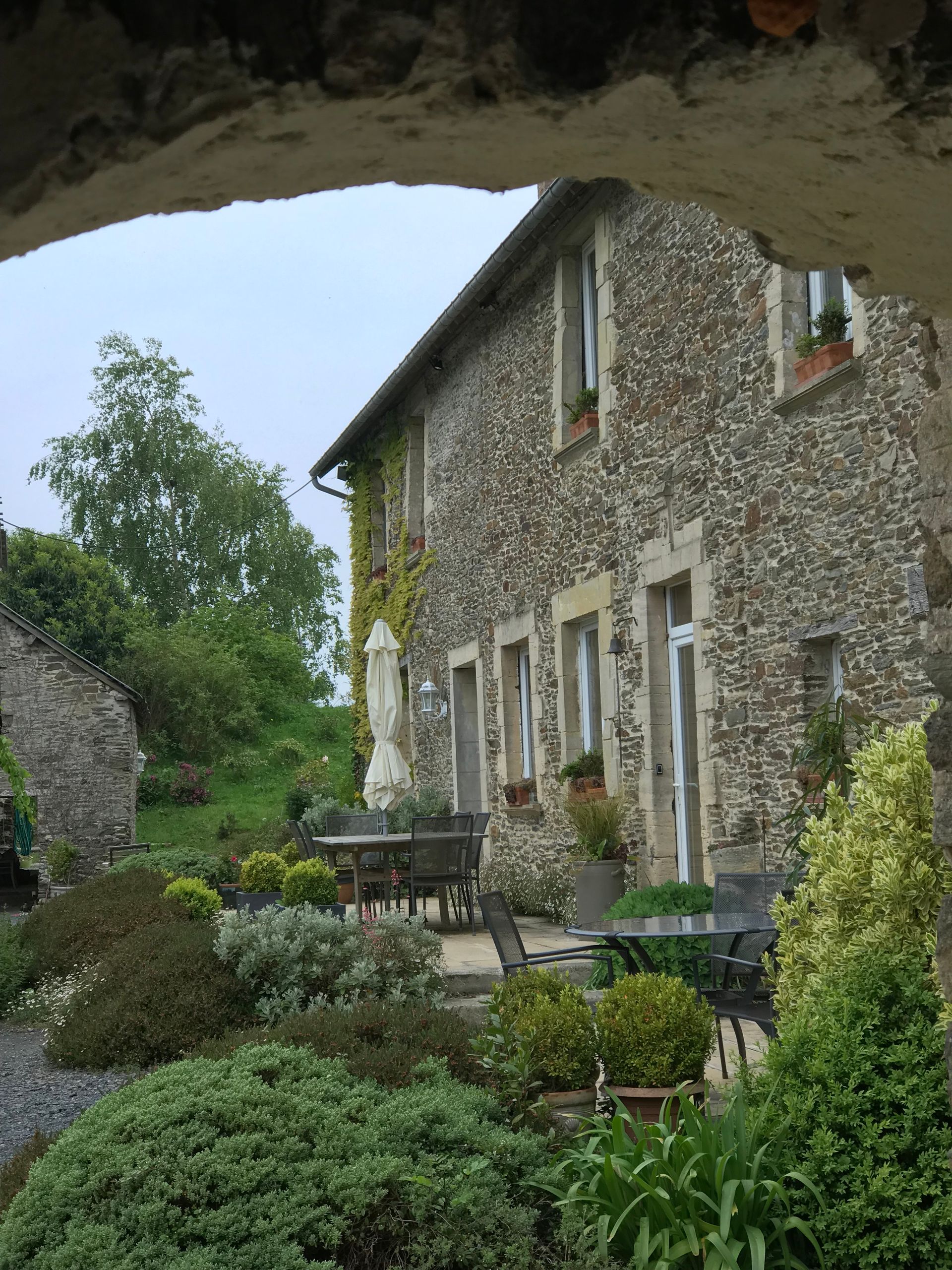 Une maison en pierre vue à travers une arche en pierre, avec un patio et un jardin luxuriant au premier plan sous un ciel nuageux.