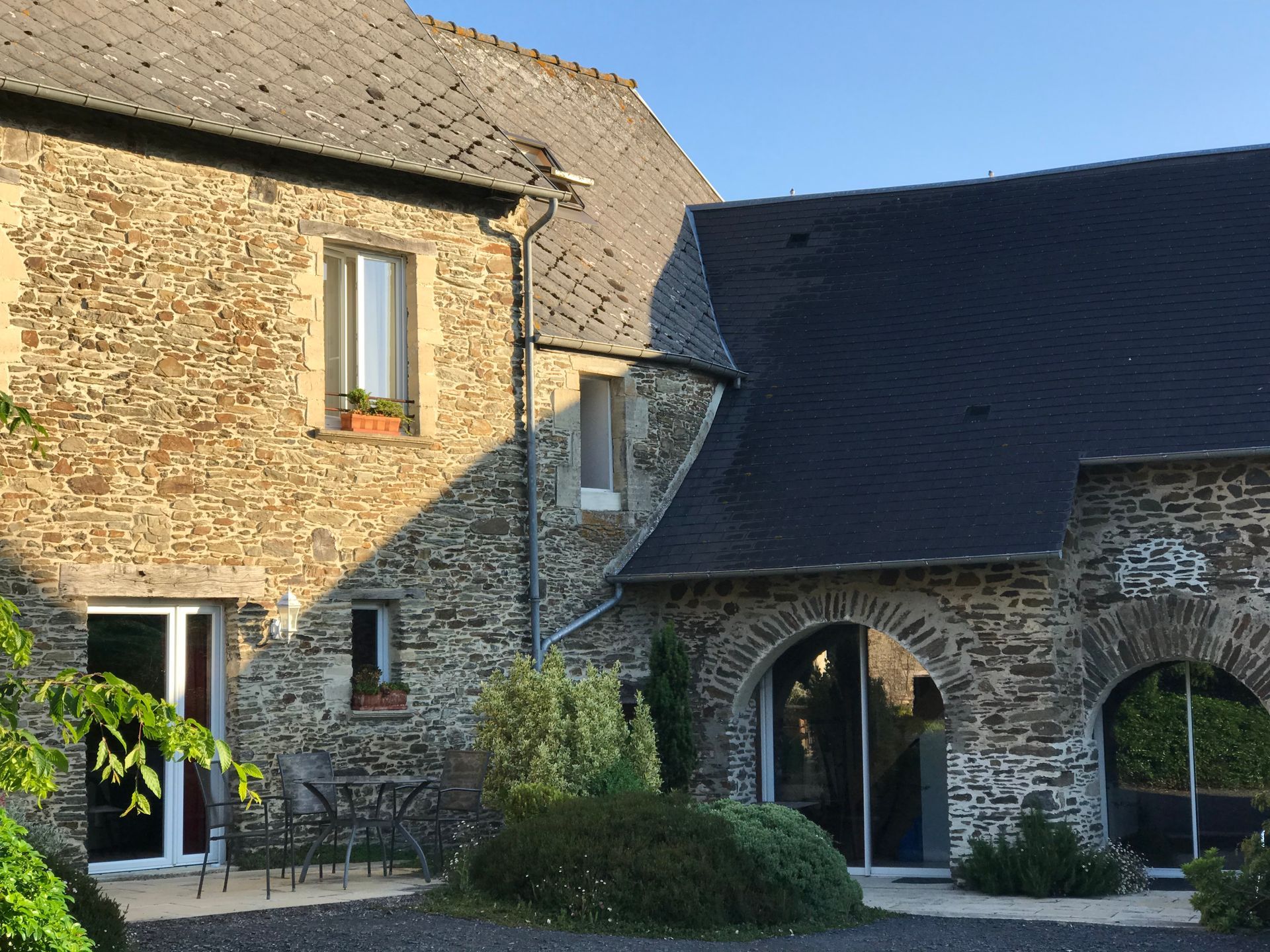 Une maison rustique en pierre, avec un toit en ardoise et de grandes portes cintrées en verre, entourée d'une cour pavée et de verdure.