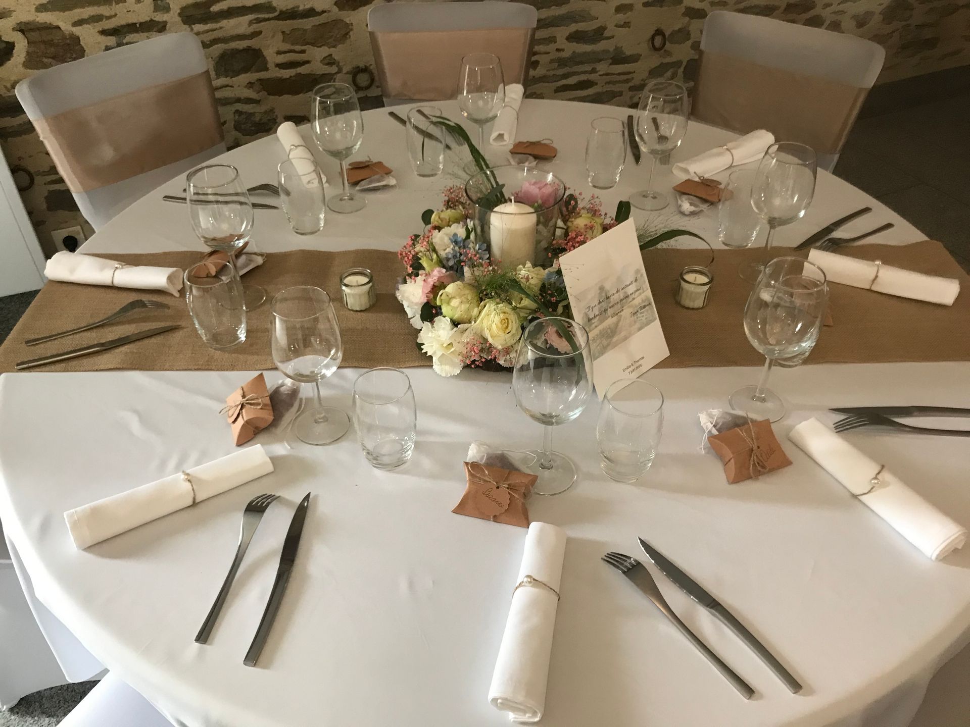 Une table de mariage ronde avec nappes blanches, chemins de table en toile de jute, un centre de table floral, des couverts et des chaises.