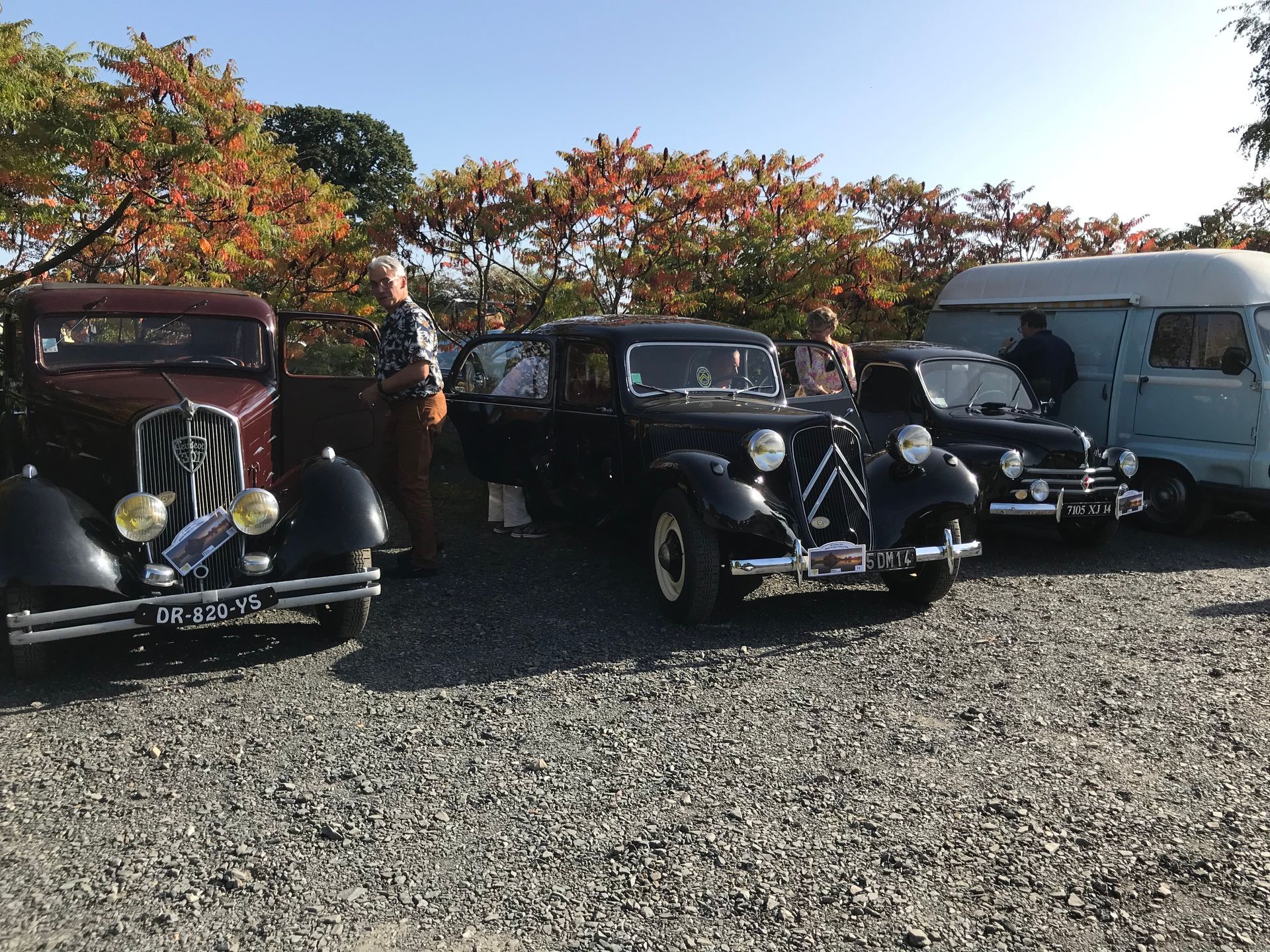 Une rangée de voitures anciennes garées sur du gravier près d'arbres aux couleurs d'automne et d'une camionnette blanche