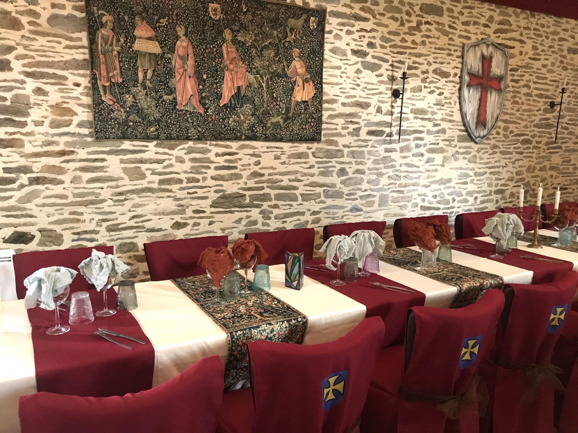 Une salle à manger de style médiéval présente de longues tables recouvertes de nappes blanches, de chemins de table bordeaux et de chaises ornées de croix dorées.